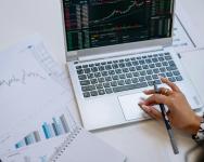 A laptop sits on a white table alongside papers with market reports on them. A woman holds a pen and one paper while using the touchpad on the laptop.