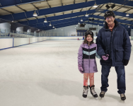 A man and a child are dressed in winter clothes and wearing skates. They are standing in an indoor skating rink. Both are smiling.
