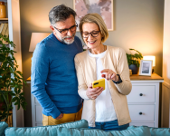 A middle-aged couple smiles while looking at a phone. They are standing in their home behind a couch.